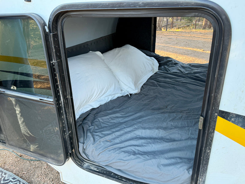 Inside an RV rental's sleeping area, showing a bed with white pillows and gray sheets. Door open to outdoors. — photo 8