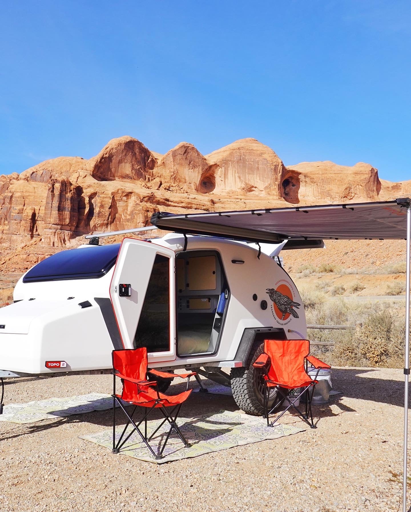 This white teardrop RV rental is set up for desert camping, with chairs and awning, backed by majestic red rock formations.
