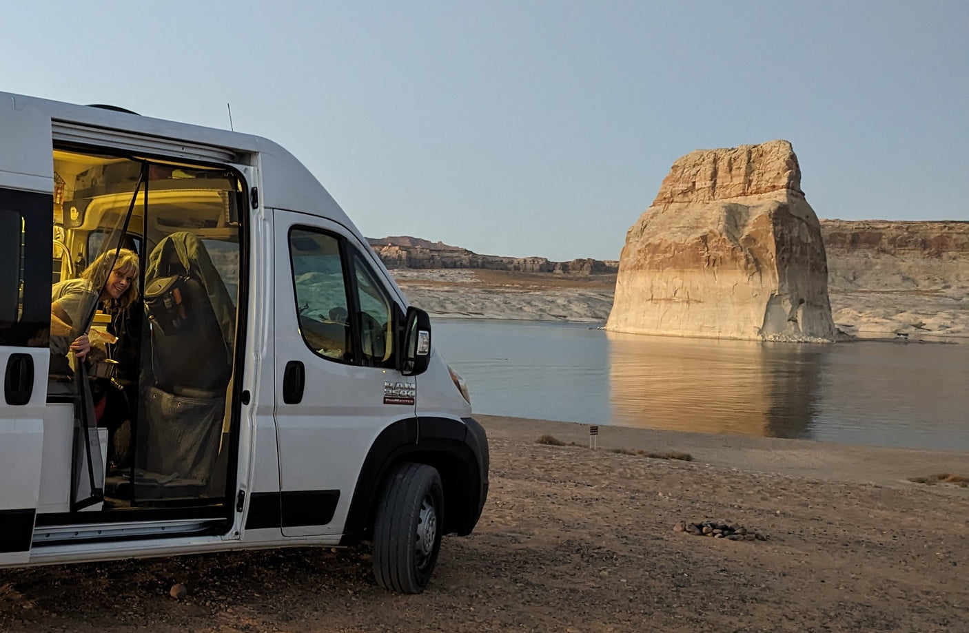RV rental trip: Woman enjoys the view from her camper van, parked on Lake Powell's shore near an illuminated rock formation.