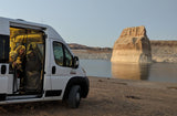 RV rental trip: Woman enjoys the view from her camper van, parked on Lake Powell's shore near an illuminated rock formation.