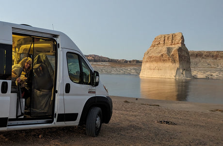 RV rental trip: Woman enjoys the view from her camper van, parked on Lake Powell's shore near an illuminated rock formation.