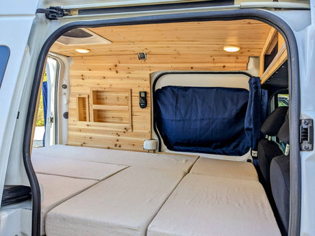 Inside a custom camper van, light wood panels line the walls and ceiling. A comfy, modular bed stretches across the floor. This inviting RV rental features built-in storage and a dark curtain.