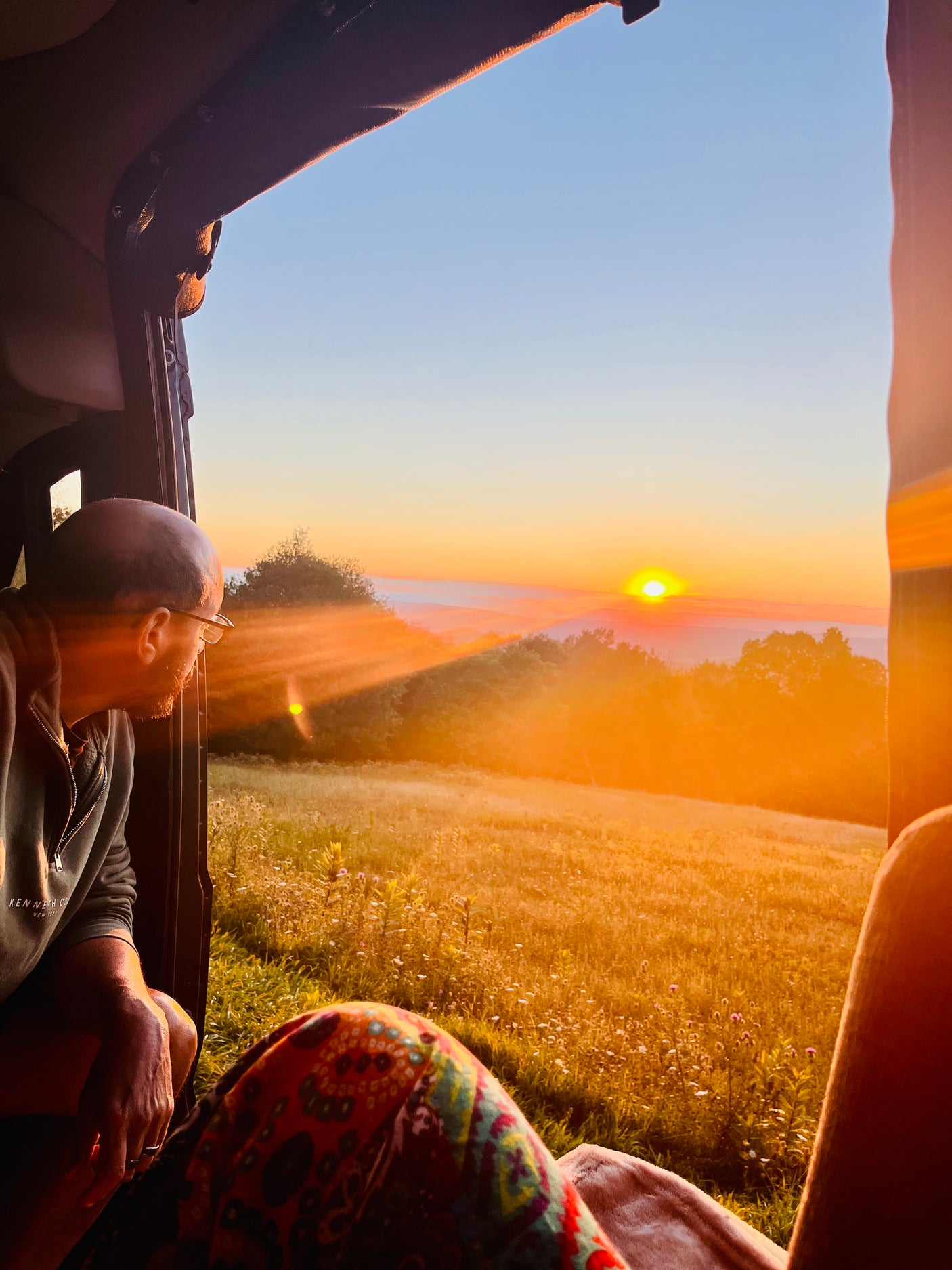 A man in an RV rental watches a vibrant sunrise over a golden field and distant mountains. Warm light floods the view.