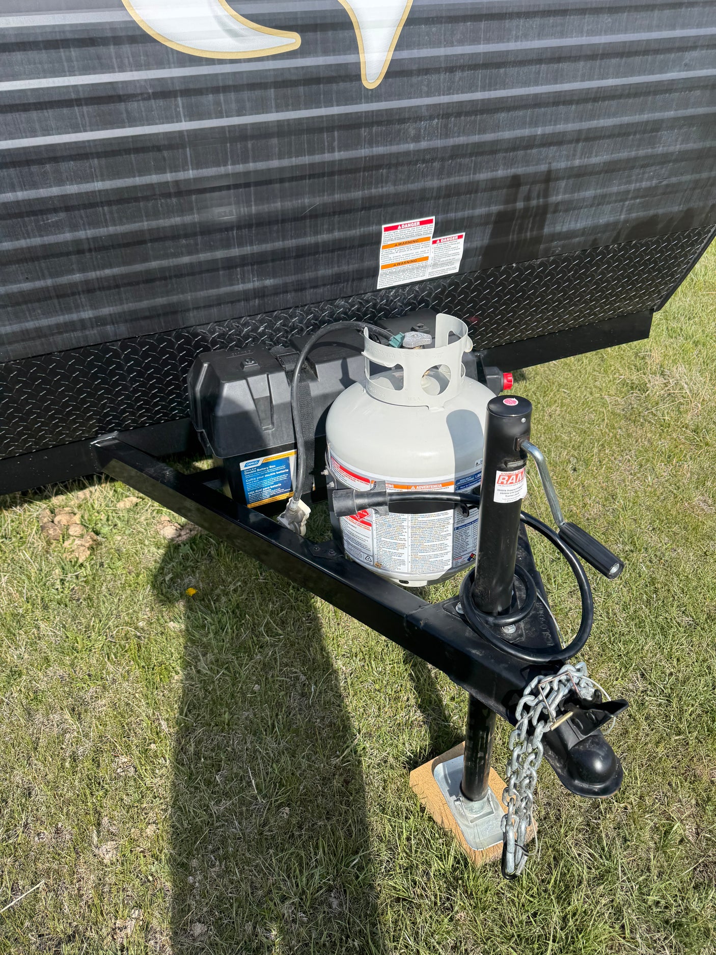 RV rental trailer's front hitch area with propane tank, CAMCO double battery box, and RAM jack on grass.