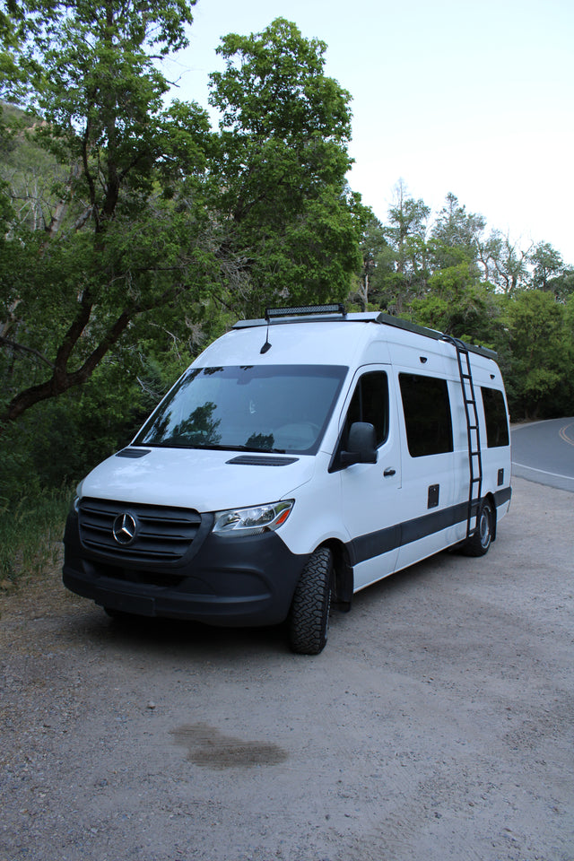 White Mercedes Sprinter rv rental camper van with a roof light bar, parked on a dirt road next to a winding paved road surrounded by trees.
