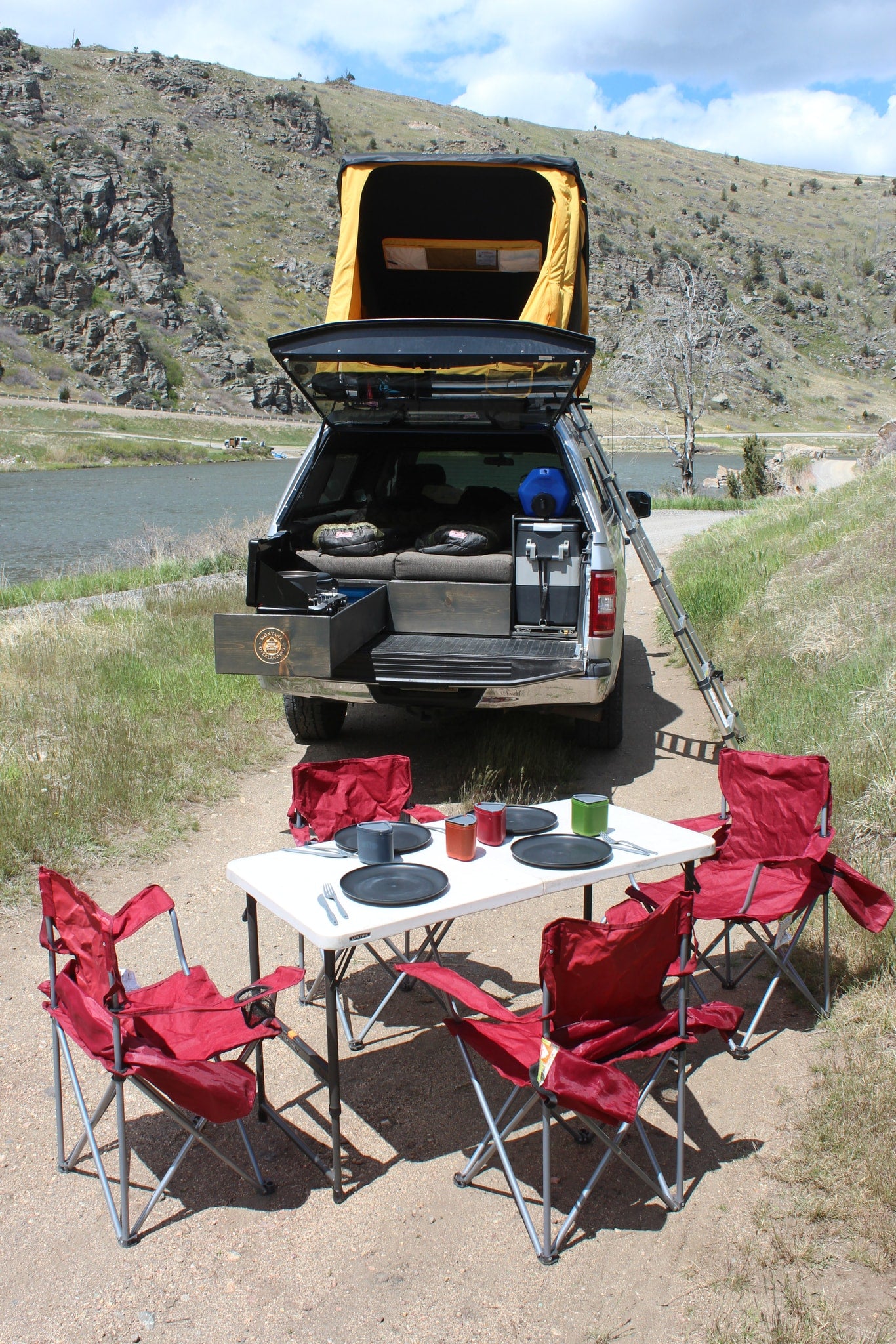 This RV rental showcases an overlanding truck camper with a rooftop tent, portable kitchen, and outdoor dining setup by the river. — photo 10