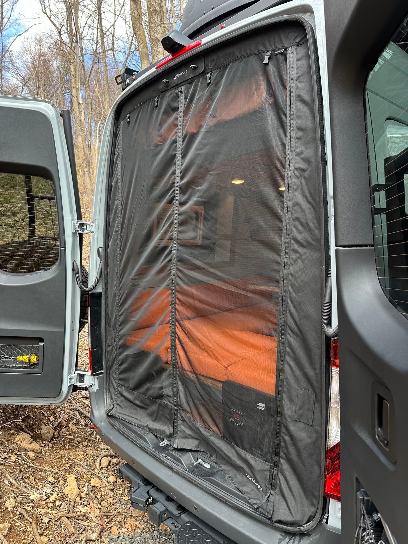 This RV rental features a VanEssential insect screen on its open rear doors, revealing a comfy interior bed for outdoor living.