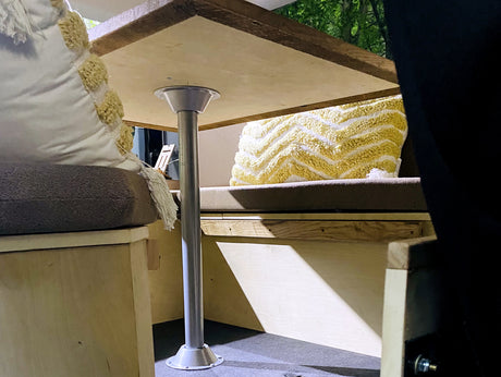 Inside an RV rental: a wooden table, cushioned benches with decorative pillows, with trees visible outside.