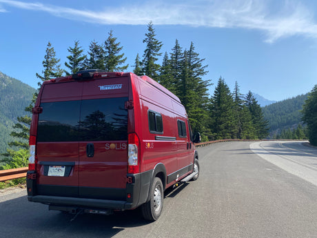 This red Winnebago Solis rv rental van sits on a winding mountain road through dense forests. Perfect for scenic trips!