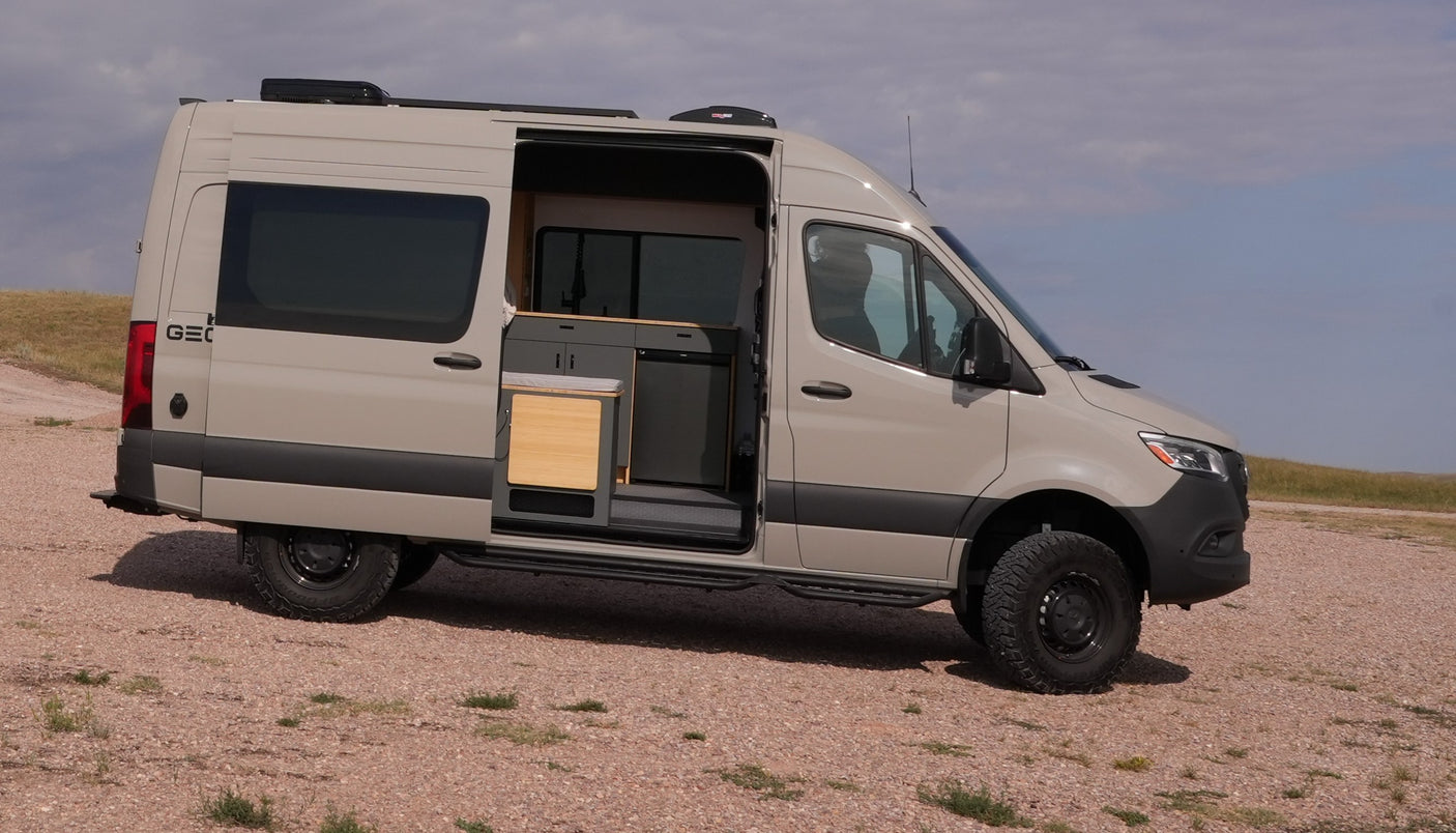 This adventure-ready light tan camper van, ideal for rv rental, sits on a gravel path, its side door open to a compact kitchen.