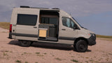This adventure-ready light tan camper van, ideal for rv rental, sits on a gravel path, its side door open to a compact kitchen.