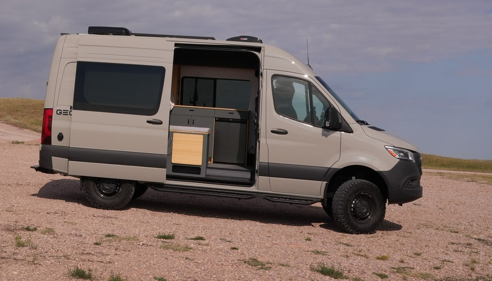 This adventure-ready light tan camper van, ideal for rv rental, sits on a gravel path, its side door open to a compact kitchen.