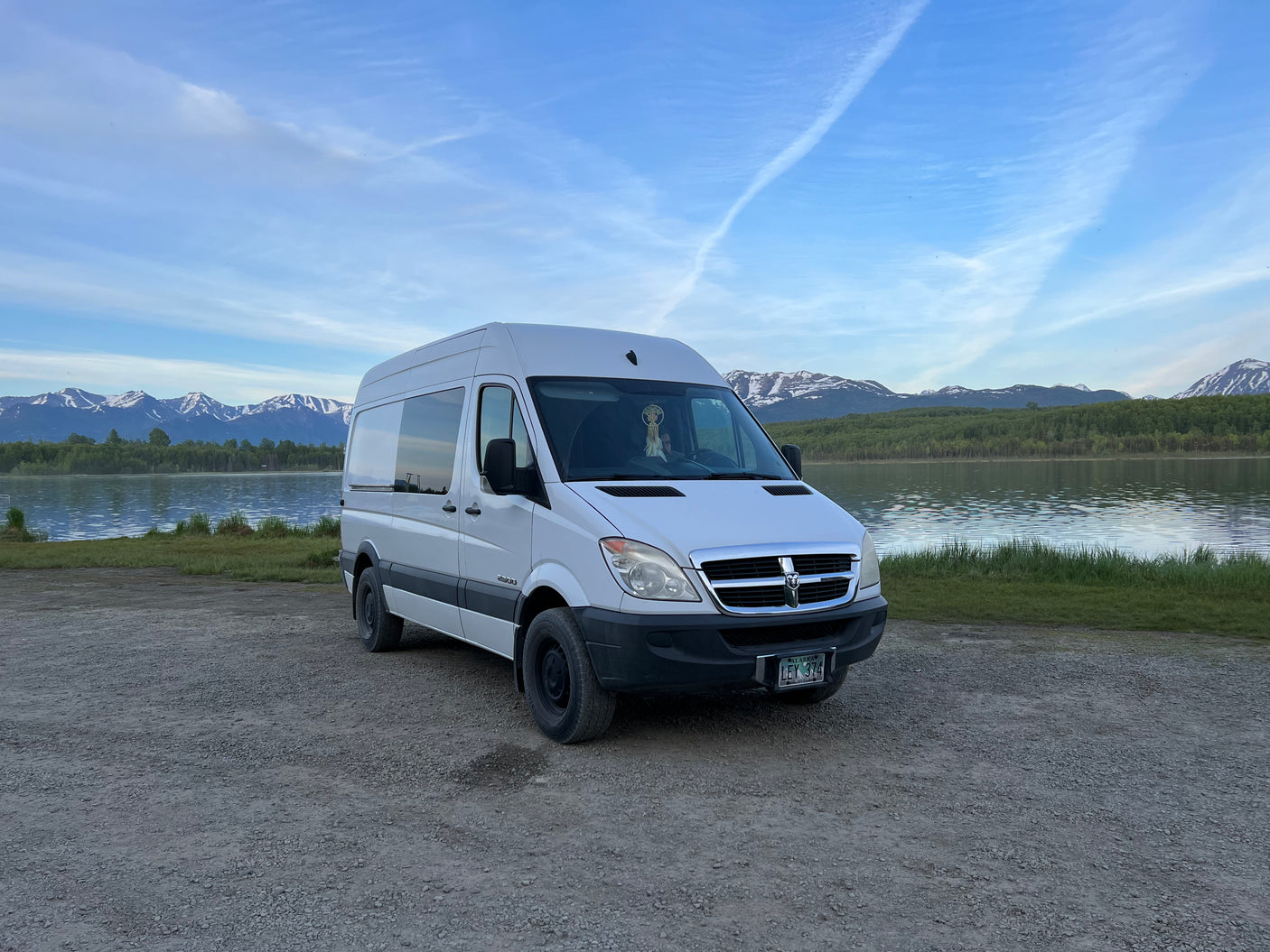 This spacious white camper van, ideal for an RV rental, is parked lakeside with majestic snow-capped mountains in Alaska.