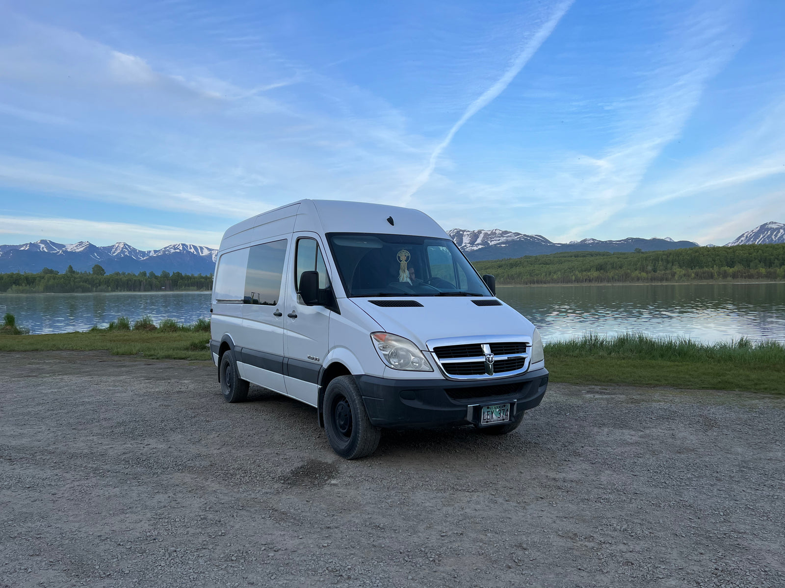 This spacious white camper van, ideal for an RV rental, is parked lakeside with majestic snow-capped mountains in Alaska.