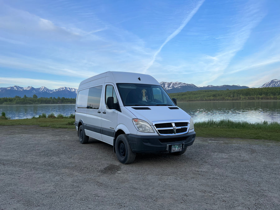 This spacious white camper van, ideal for an RV rental, is parked lakeside with majestic snow-capped mountains in Alaska.
