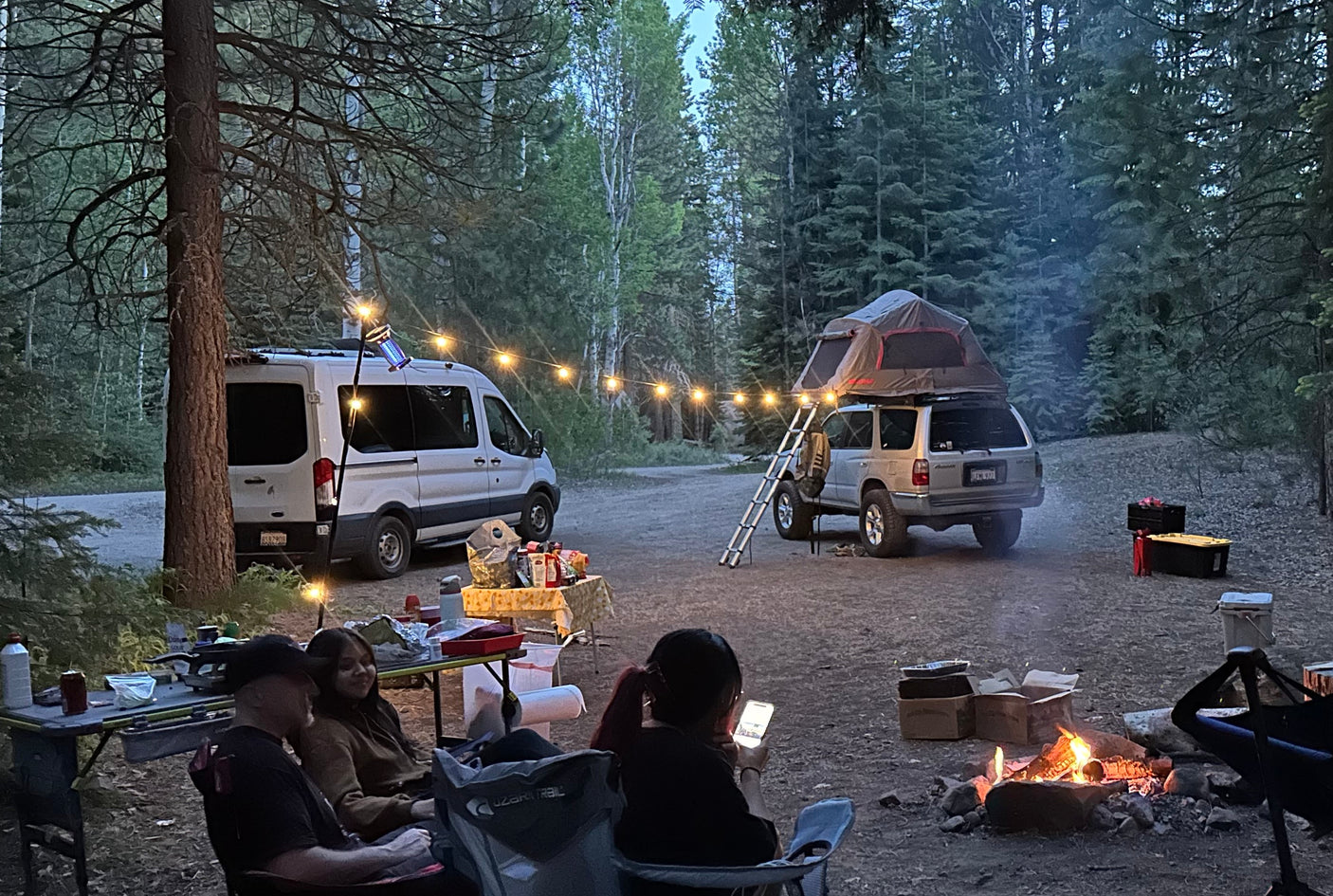 Campsite fun with an RV rental van, SUV with rooftop tent, campers around a fire, and string lights at dusk.