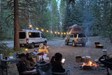 Campsite fun with an RV rental van, SUV with rooftop tent, campers around a fire, and string lights at dusk.