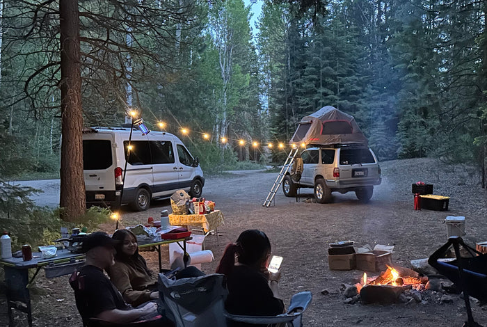 Campsite fun with an RV rental van, SUV with rooftop tent, campers around a fire, and string lights at dusk.