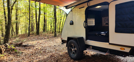 Enjoy nature! This cozy teardrop RV rental, parked in a vibrant forest, welcomes you with an open door to a bed and AC.