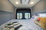 An RV rental's cozy bed with patterned linens & adventure pillows, looking out back doors onto a paved area.