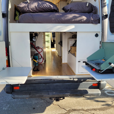 Inside an RV rental: a raised bed, under-bed storage with power and water systems, plus a rear outdoor kitchen with a portable stove.