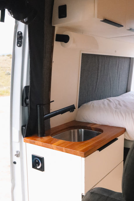 Inside this rv rental, a modern sink, wooden counter, and functional control panel are next to a cozy bed.