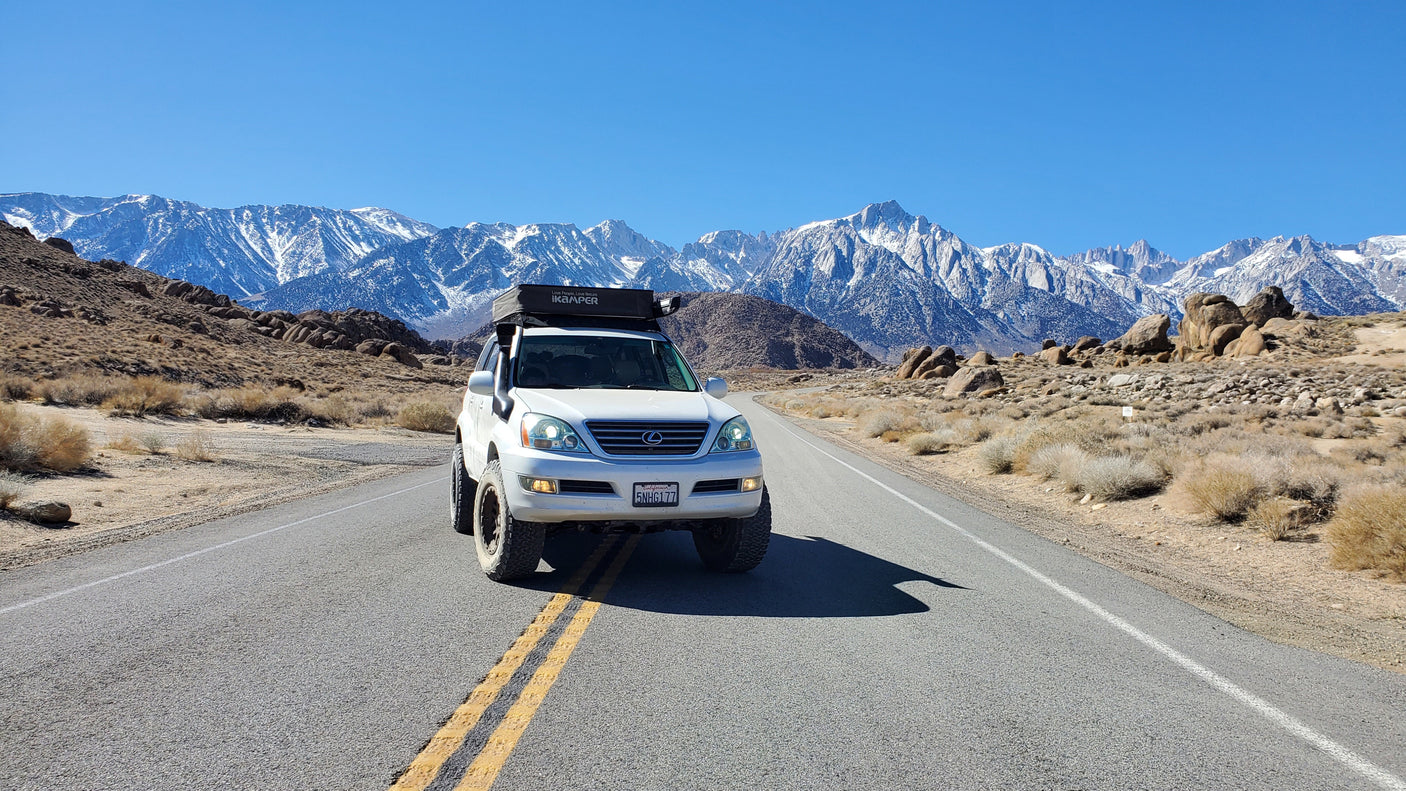 White Lexus GX with rooftop tent on a desert road, snow-capped mountains. A unique choice for your RV rental adventure.