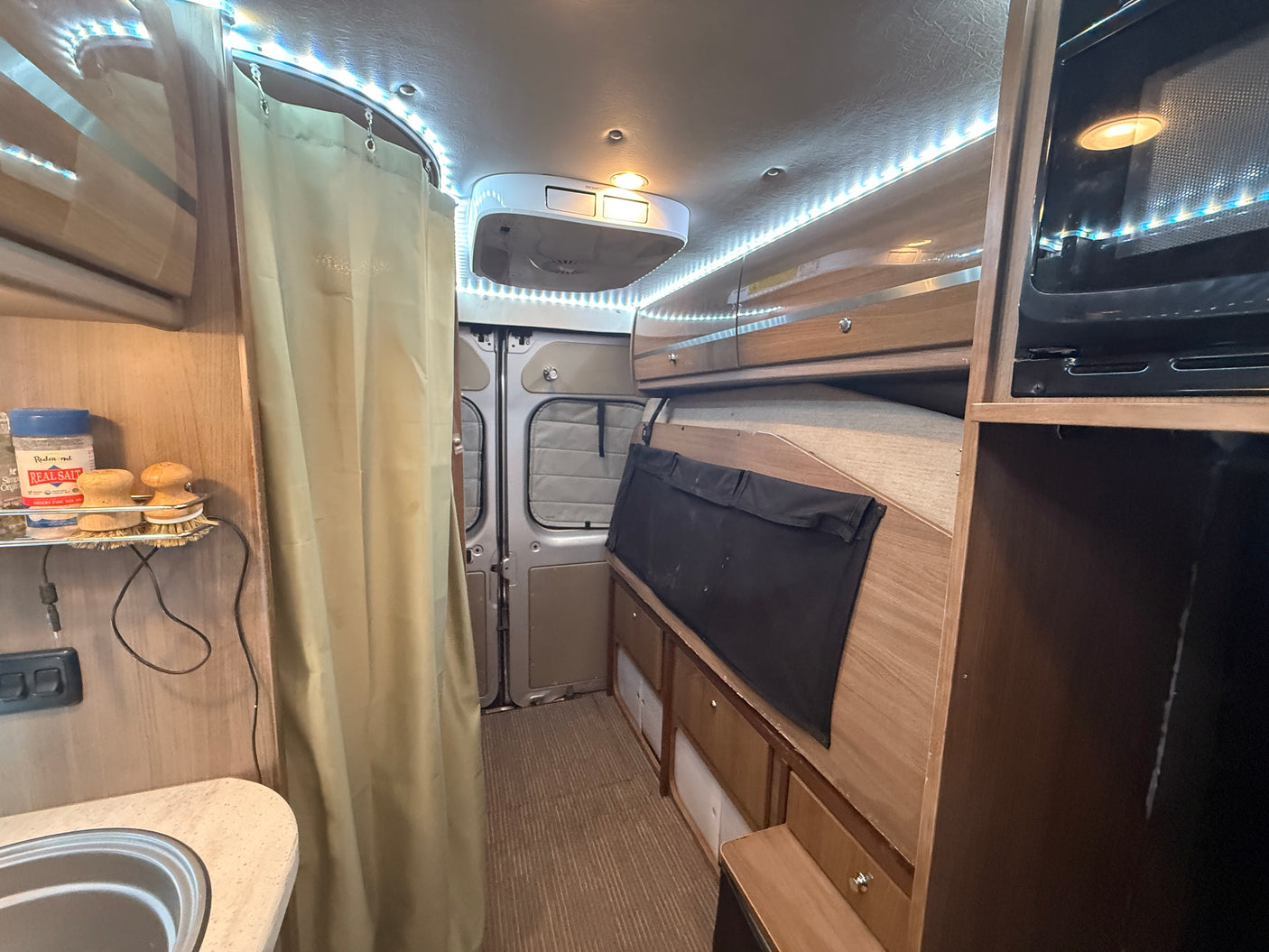 An RV rental interior showing a compact kitchen with sink, curtained area, rear van doors, cabinetry, and bright overhead lighting.