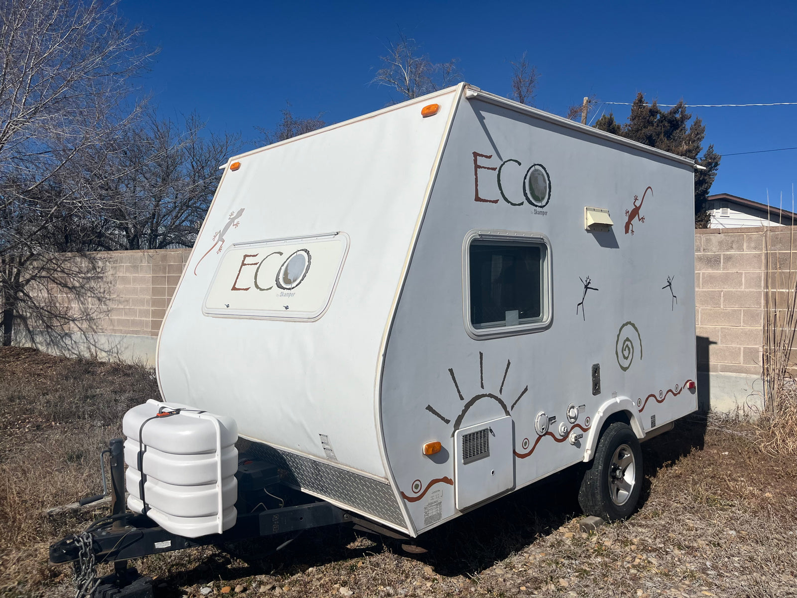 White Eco Skamper RV rental: travel trailer with lizard & sun decals, parked outdoors under blue sky.