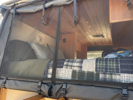 View into a cozy RV rental bedroom. A bed with plaid quilt, wood ceiling, & fan are visible through a protective mesh.