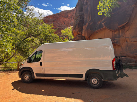 White cargo-style RV rental van with a bike rack, parked in a scenic desert canyon among green trees. Perfect for adventure travel.