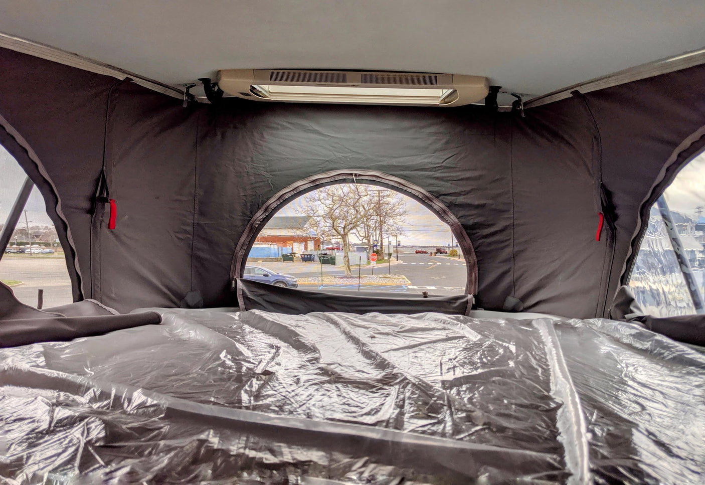 Interior of an RV rental pop-top, with a plastic-covered sleeping area, offering an outdoor view of a street and bare trees.