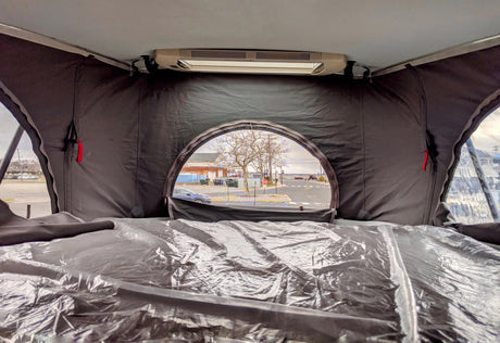 Interior of an RV rental pop-top, with a plastic-covered sleeping area, offering an outdoor view of a street and bare trees.