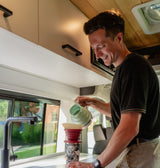 Smiling man makes pour-over coffee in an RV rental. He brews a fresh cup with a travel mug inside the modern camper van.
