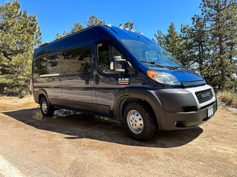 Blue Ram ProMaster RV rental van on a dirt road with puddles & pine trees under a clear sky. Your next adventure awaits!|A happy older black dog sits on a rug inside a customized RV rental — photo 1