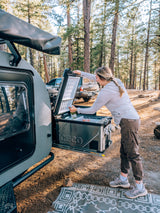 Woman restocking ICECO fridge on an RV rental teardrop camper in a sunny pine forest. Ready for adventure!