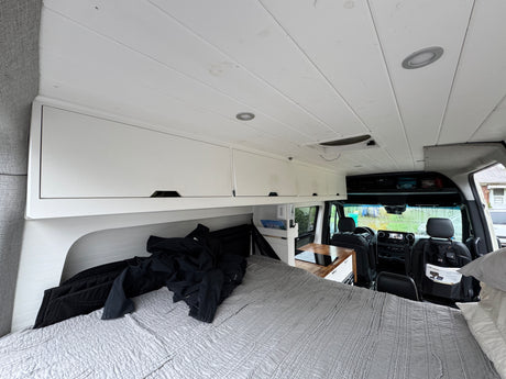 View from the back of an RV rental, showing a grey bed, white overhead cabinets, and a compact kitchen counter, leading to the front cab.