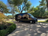 Sleek black Mercedes Sprinter van, an RV rental, parked on a sunny driveway, reflecting homes and trees on its glossy surface.