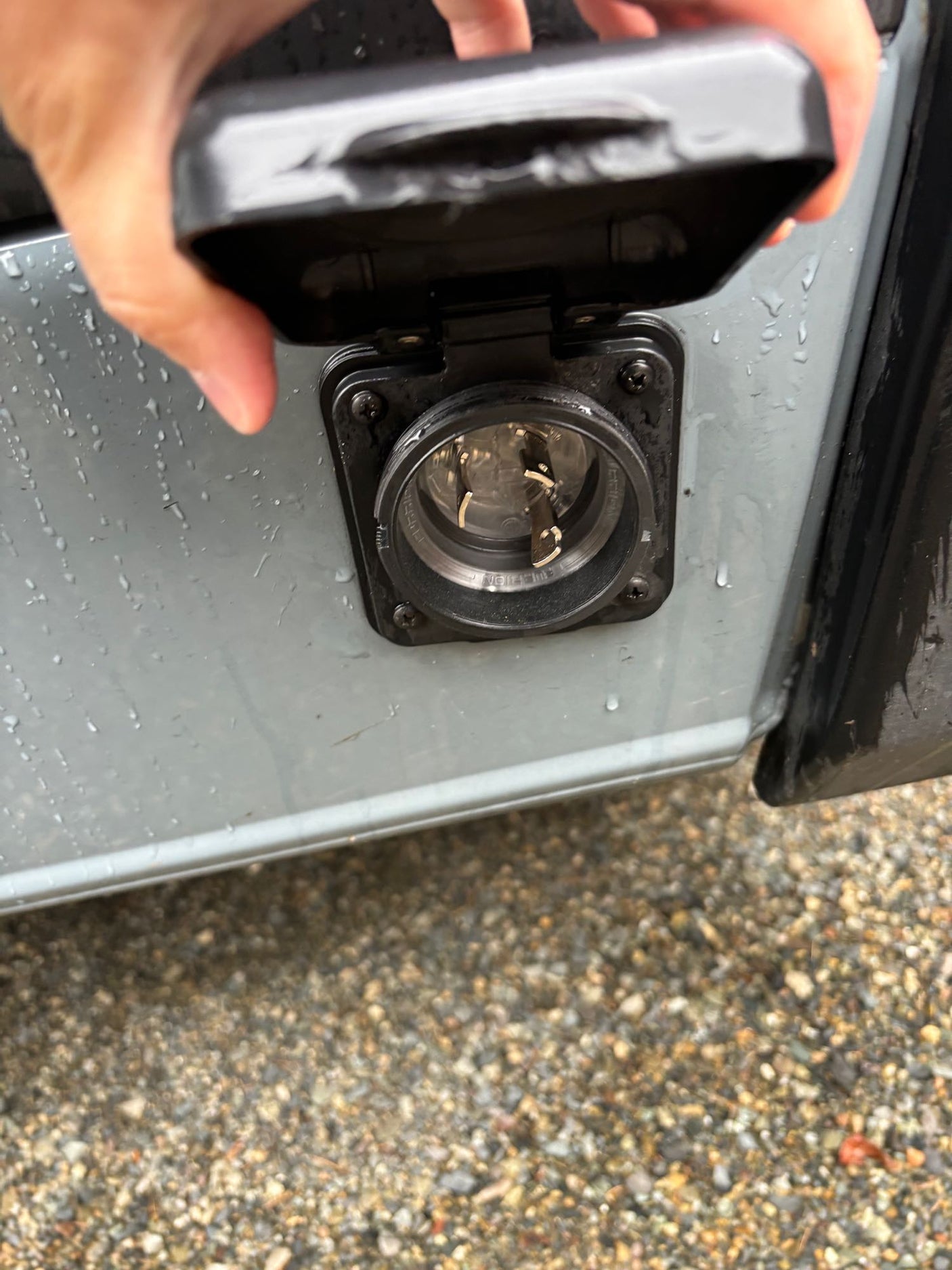 RV rental power inlet. A hand lifts the cover of a wet external electrical port on a grey RV, revealing connection prongs.