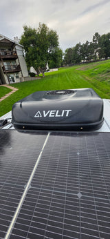 RV rental rooftop: AVELIT air conditioner and solar panels prominent. Background features an apartment building, green grass, and trees.