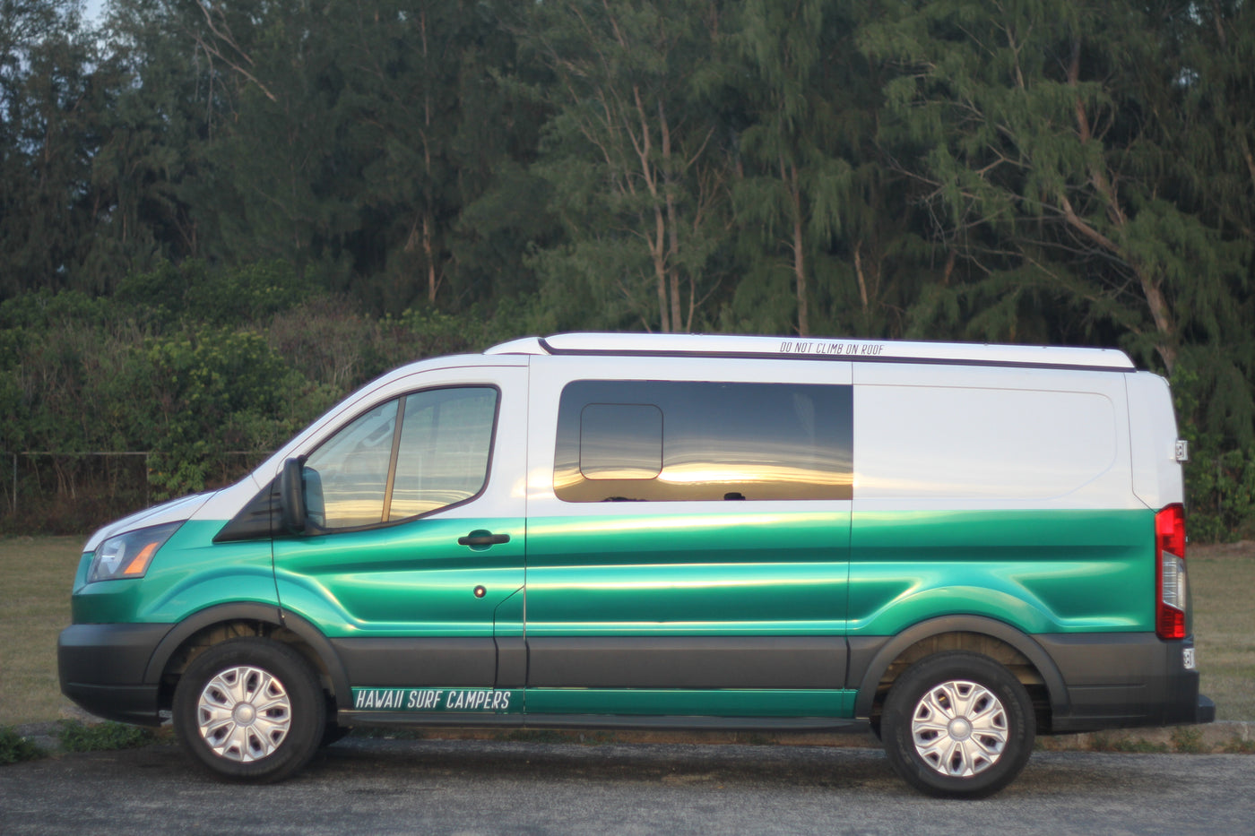 Side view of a green & white Hawaii Surf Campers RV rental van with "DO NOT CLIMB ON ROOF" visible, reflecting the sunset.