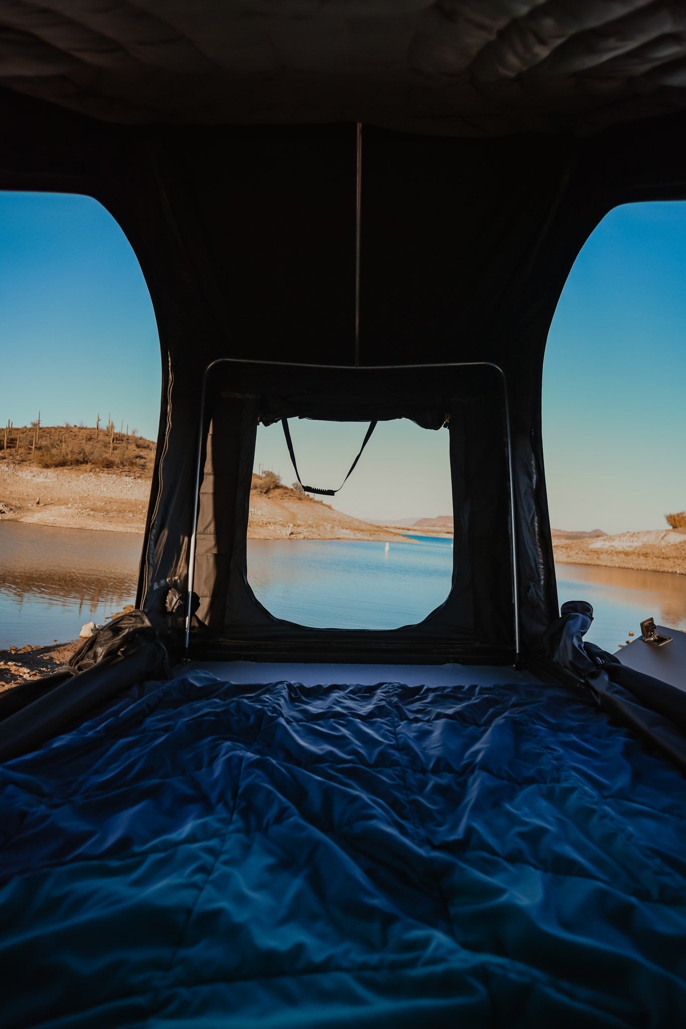 Cozy rv rental rooftop tent with a blue comforter, offering serene views of a desert lake and distant cacti. — photo 20