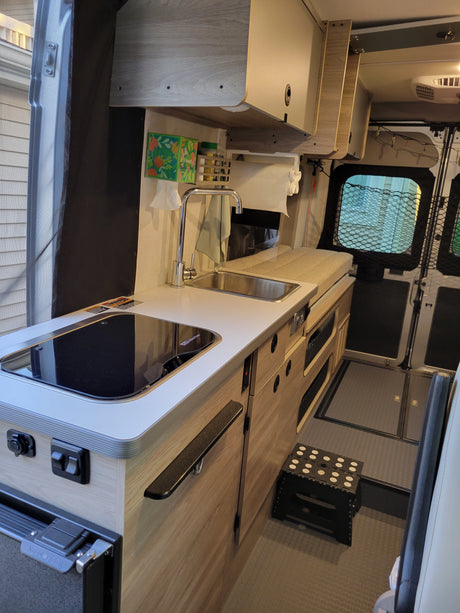 Inside an RV rental, showing a compact kitchen with an induction cooktop, sink, fridge, & storage cabinets.
