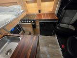RV rental interior: cozy bed, compact kitchen with stove, sink, fridge, wooden counters, and a Bluetti power station.
