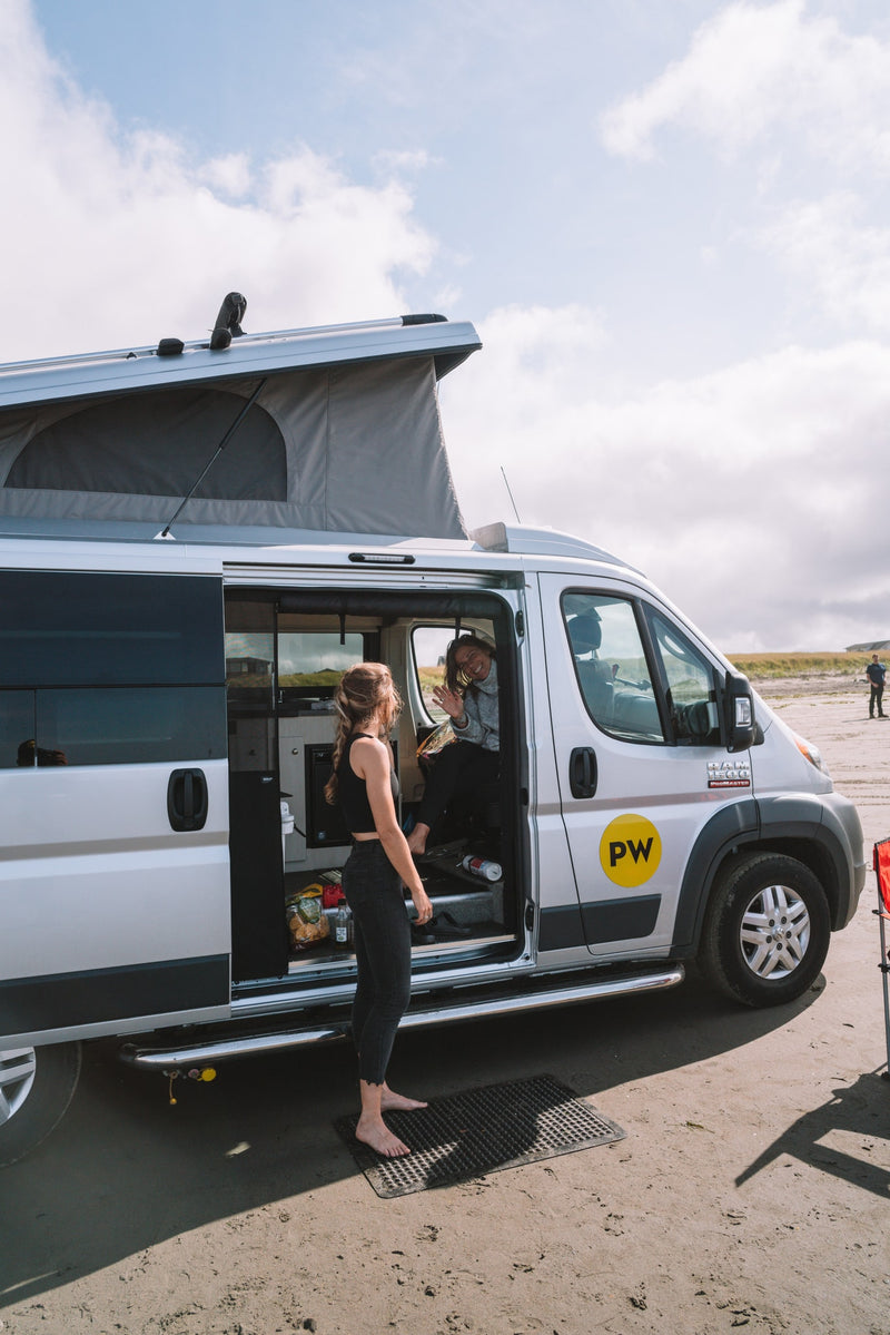 Two women by their pop-top rv rental camper van on a sandy spot. One stands barefoot outside, another waves from inside. — photo 7