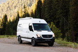 A white Wasatch custom Sprinter rv rental camper van, parked on a gravel road amidst a vibrant mountain forest. Your next adventure awaits!