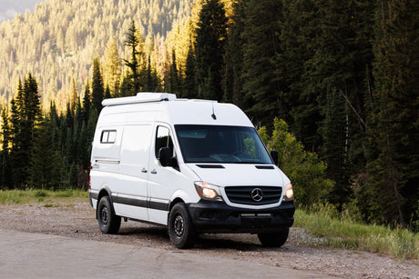 A white Wasatch custom Sprinter rv rental camper van, parked on a gravel road amidst a vibrant mountain forest. Your next adventure awaits!