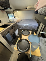 Cozy RV rental interior: kitchen on left, bed & bunk in back, dining setup with black/blue dishes in foreground.