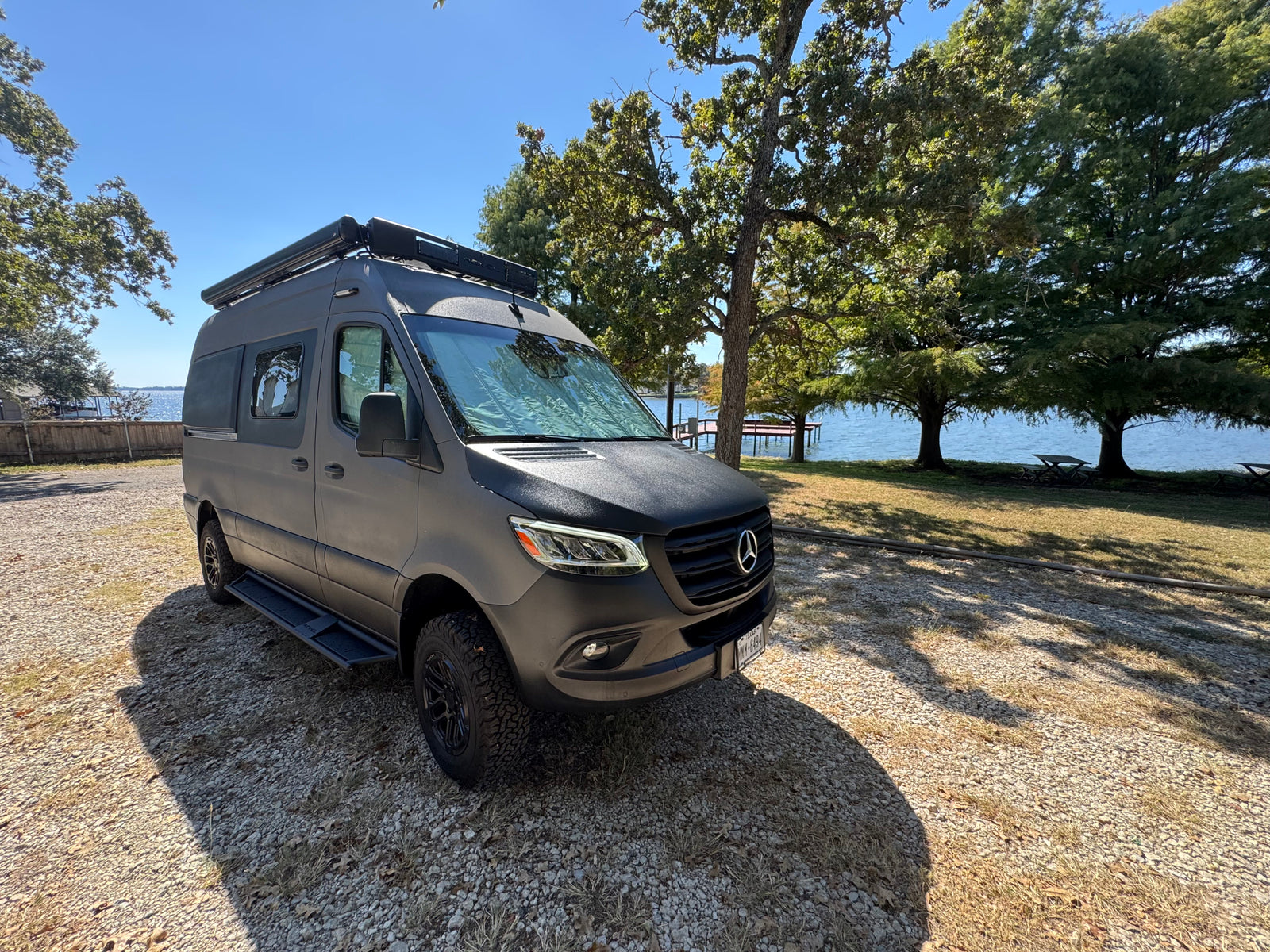 Black Mercedes Sprinter RV rental parked on a gravel path by a scenic lake and trees, perfect for a van life adventure.