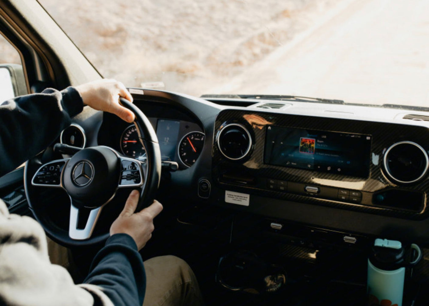 Driving an RV rental: View from driver's seat. Hands on Mercedes wheel, dashboard, and infotainment system with music.